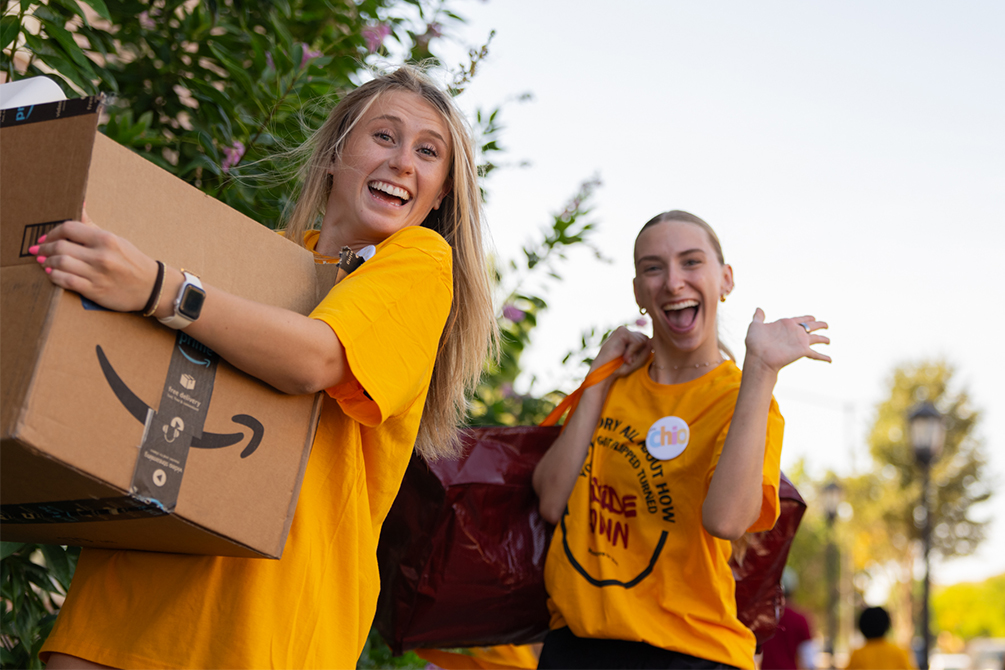 Two young ladies in gold t-shirts carry items on Move-In Day