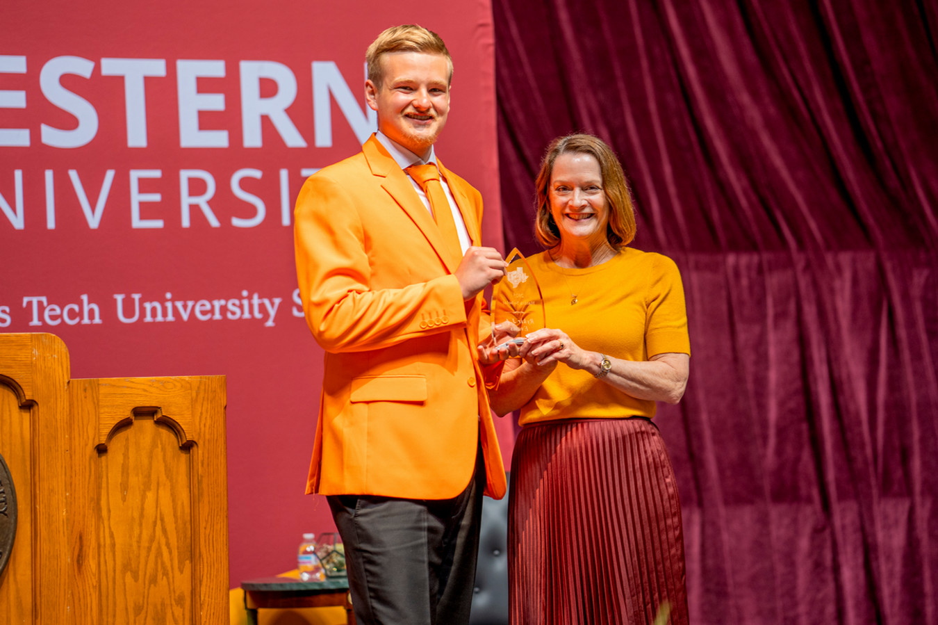 Rykir Evans in an orange suit as he accepts Clark Scholar award from Stacia Haynie